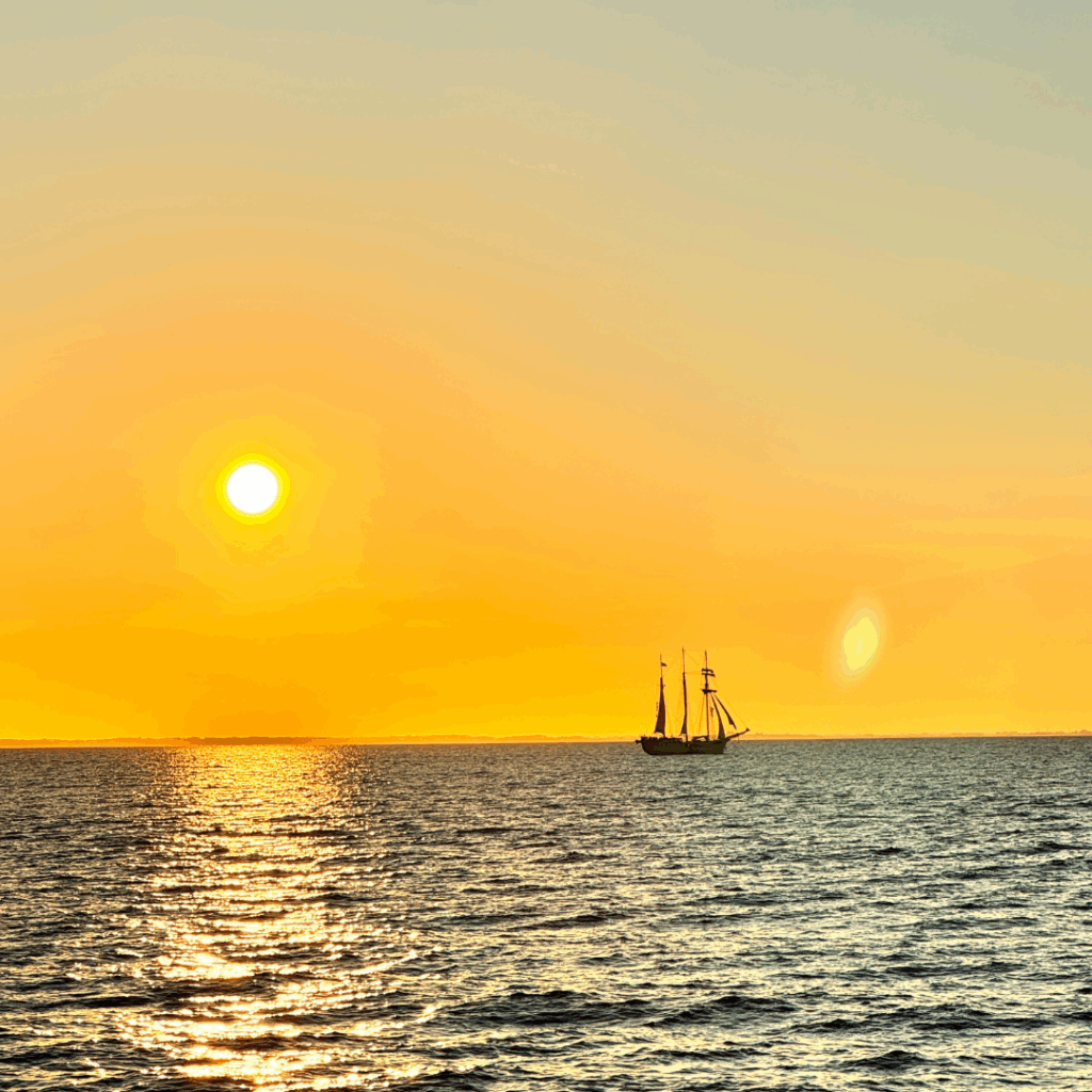 Auf dem Meer selgelt ein Boot, im Hintergrund geht die Sonne unter